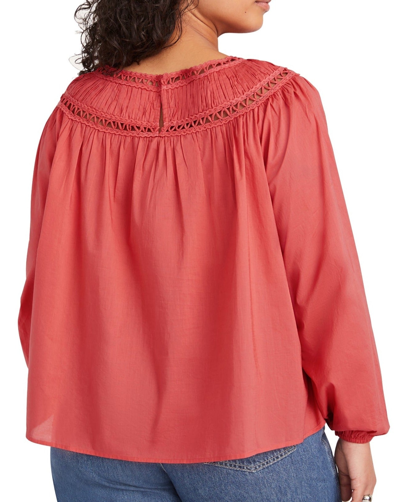 A person with curly hair is wearing a Merlette New York Merlette Ardent Top, a coral-colored, long-sleeved blouse with lace detailing on the back yoke and voluminous sleeves, paired with blue jeans.