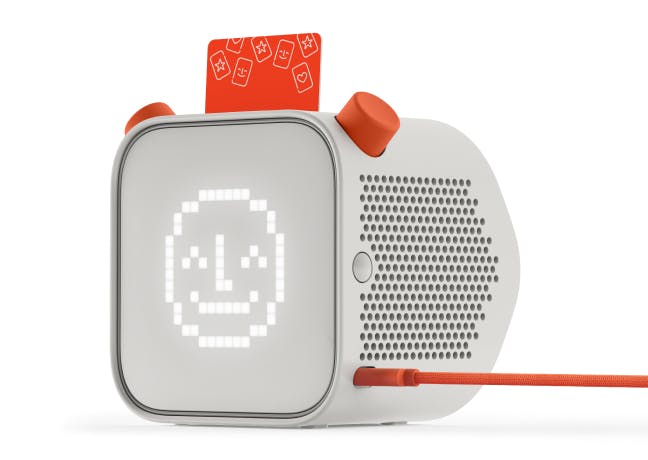 The Yoto Player by Yoto is a child-friendly audio device designed as a white cube with a pixelated face display and orange buttons. An orange card slot is located on the top, and an orange cable connects to the side, providing an ideal screen-free listening experience for kids.