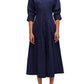 A woman stands facing the camera, wearing a dark blue, midi length Simkhai Signature Jazz dress with a collar and rolled-up sleeves, paired with black heeled sandals by Simkhai.