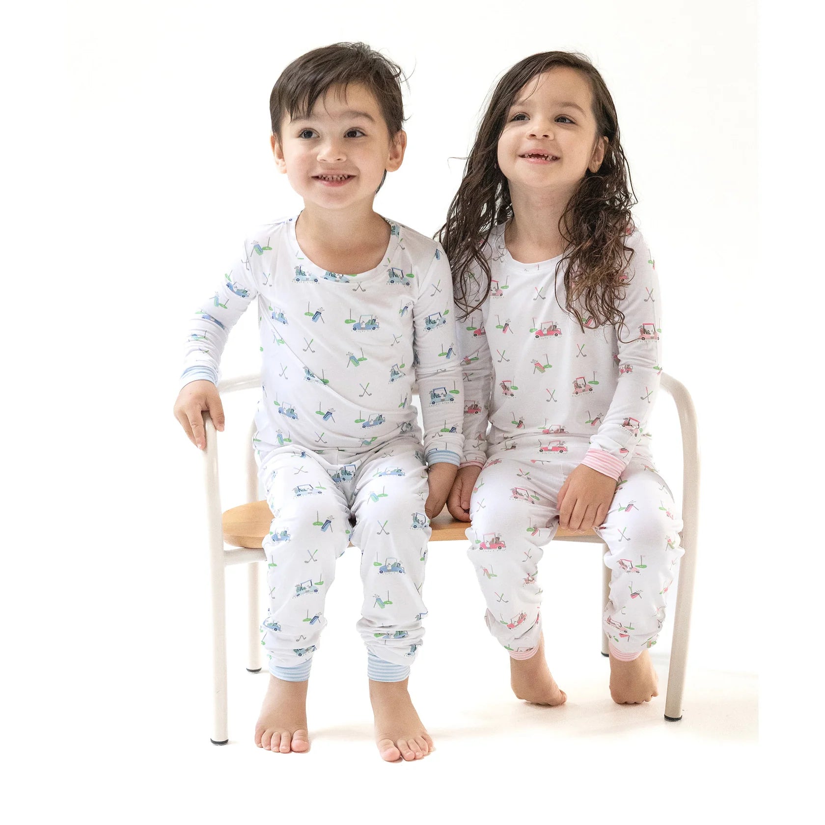 Two smiling children sit on a wooden bench, holding hands and wearing matching Angel Dear Baby and Kids' Golf Carts Long Sleeve Loungewear Sets, crafted from breathable fabric for cozy, all-day comfort.