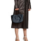 A person in a long black leather coat holds the Proenza Schouler Small Ruched Crossbody Tote while wearing white high-heeled sandals.