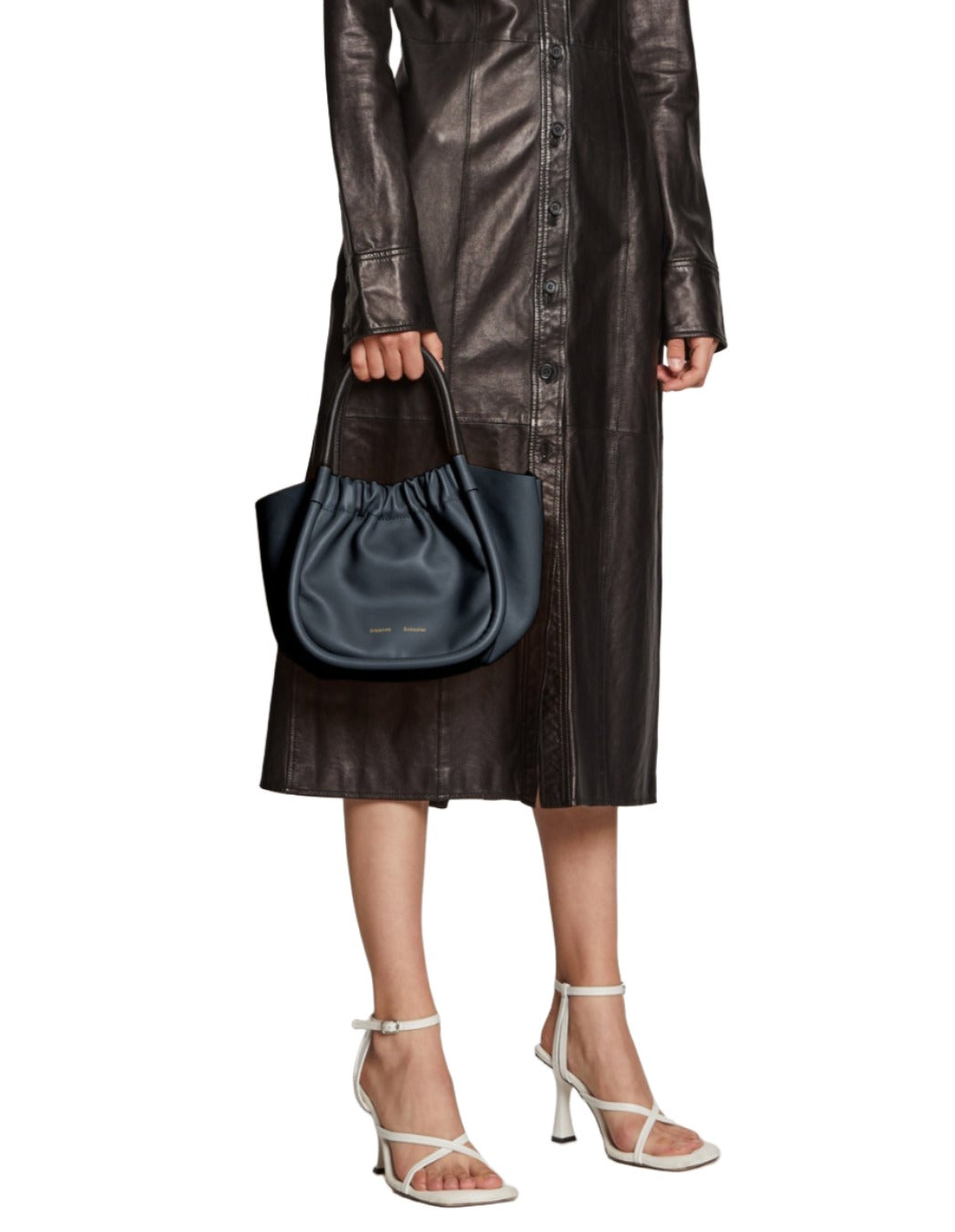 A person in a long black leather coat holds the Proenza Schouler Small Ruched Crossbody Tote while wearing white high-heeled sandals.