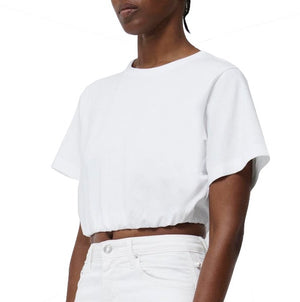 A person is wearing the Simkhai Jojo Cropped T Shirt with short sleeves and white pants, showcasing a modern style. The versatile design and relaxed fit of this Simkhai piece make the outfit perfect for any occasion. The background is plain and white.