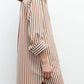 A woman stands sideways against a plain white background, wearing the TWP Morning After Dress—a brown and white vertical striped shirt dress with rolled-up sleeves and a relaxed silhouette.