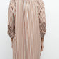 A person wears the TWP Morning After Dress, a long-sleeve brown and white vertically striped style with a relaxed silhouette and rolled-up sleeves, viewed from the back and styled with cowboy boots.