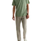 A man wearing a light green polo, AG Jeans Tellis Commuter Performance Airluxe Modern Slim beige pants, white sneakers, and a wristwatch stands against a plain white background.