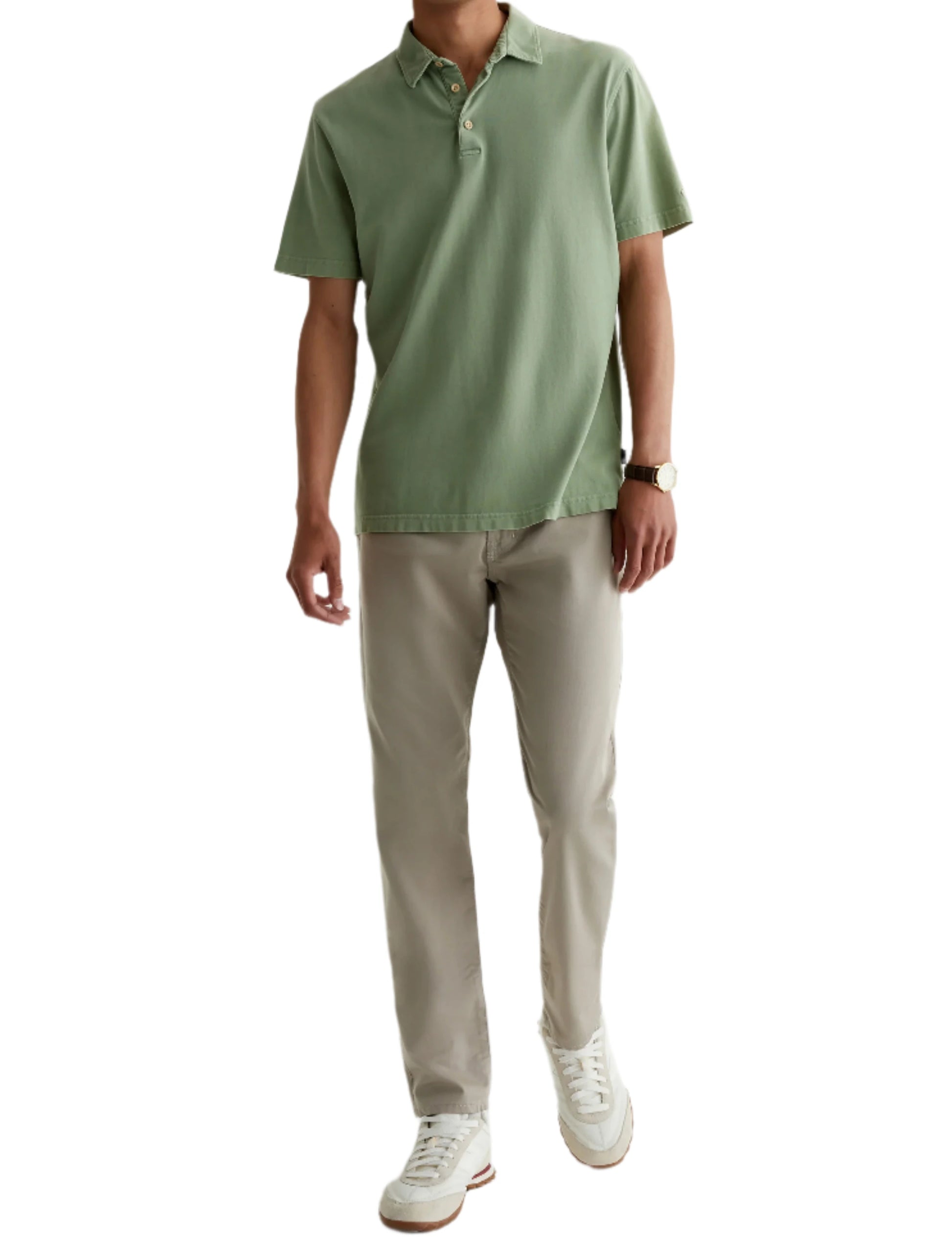 A man wearing a light green polo, AG Jeans Tellis Commuter Performance Airluxe Modern Slim beige pants, white sneakers, and a wristwatch stands against a plain white background.