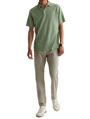 A man wearing a light green polo, AG Jeans Tellis Commuter Performance Airluxe Modern Slim beige pants, white sneakers, and a wristwatch stands against a plain white background.