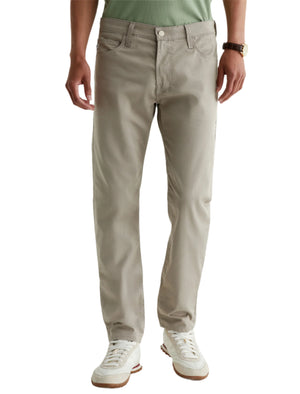 A person is shown from shoulders to feet against a plain background, wearing AG Jeans Tellis Commuter Performance Airluxe Modern Slim pants in light beige, paired with a green shirt, white sneakers, and a wristwatch.
