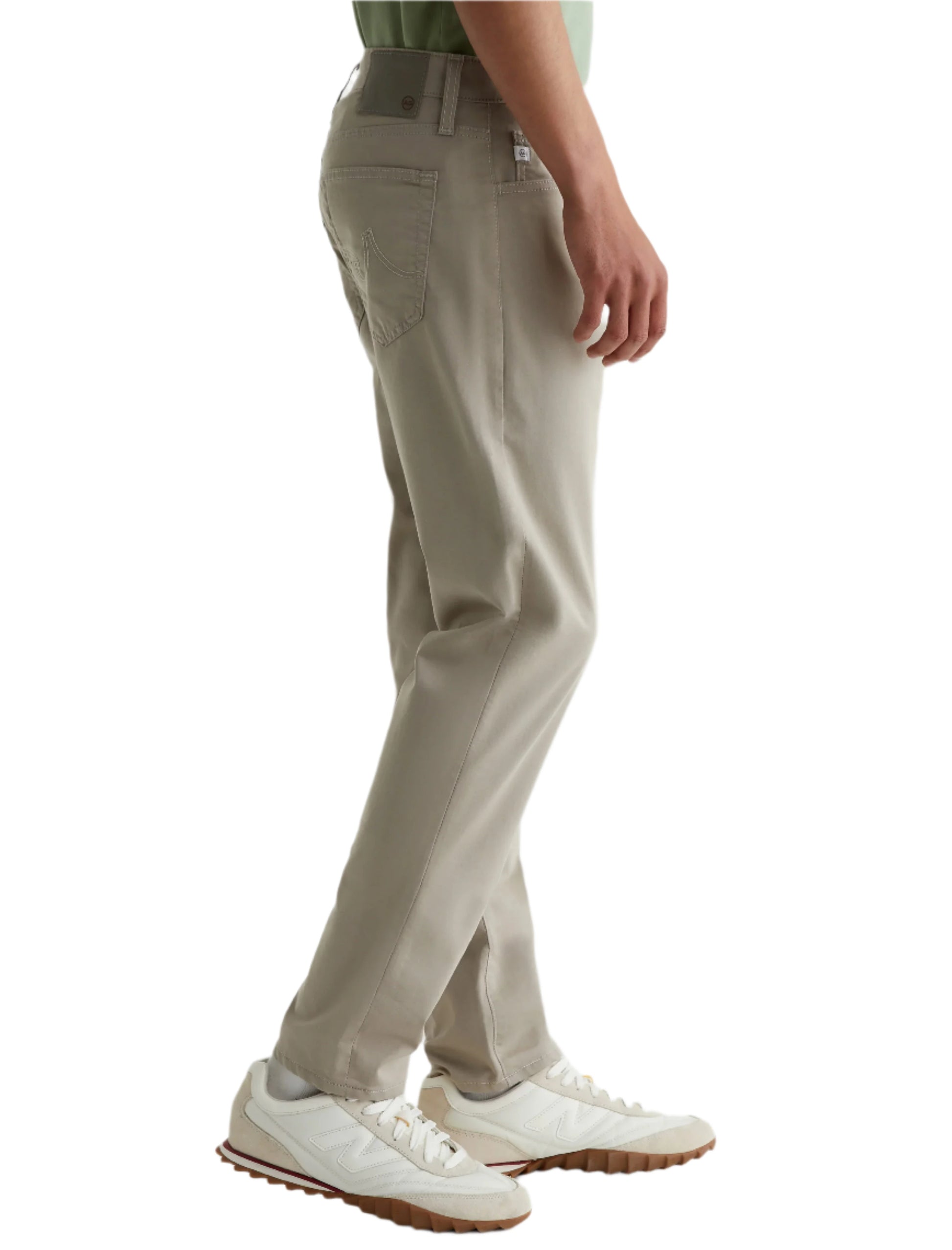 A person wearing AG Jeans Tellis Commuter Performance Airluxe Modern Slim pants in light beige, featuring a tapered leg, paired with white sneakers, is shown from the side against a plain white background.