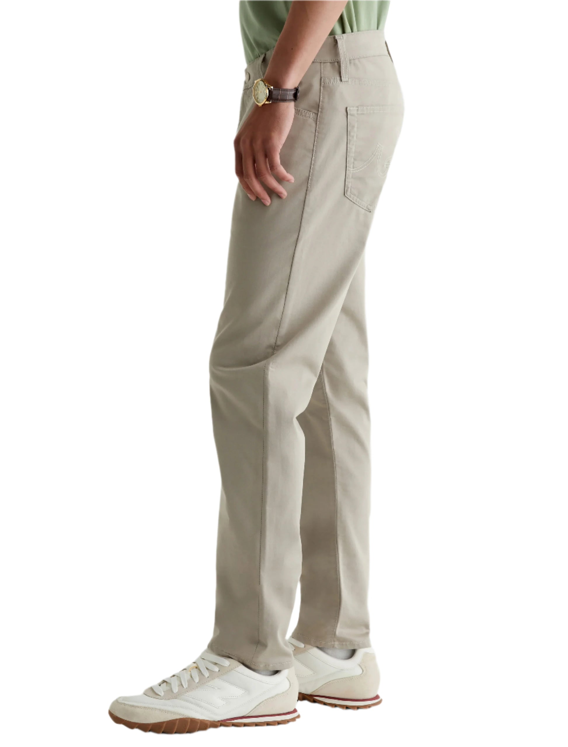 A person in AG Jeans' Tellis Commuter Performance Airluxe Modern Slim pants (beige, tapered), a green shirt, white sneakers, and a watch stands with their left side facing the camera.