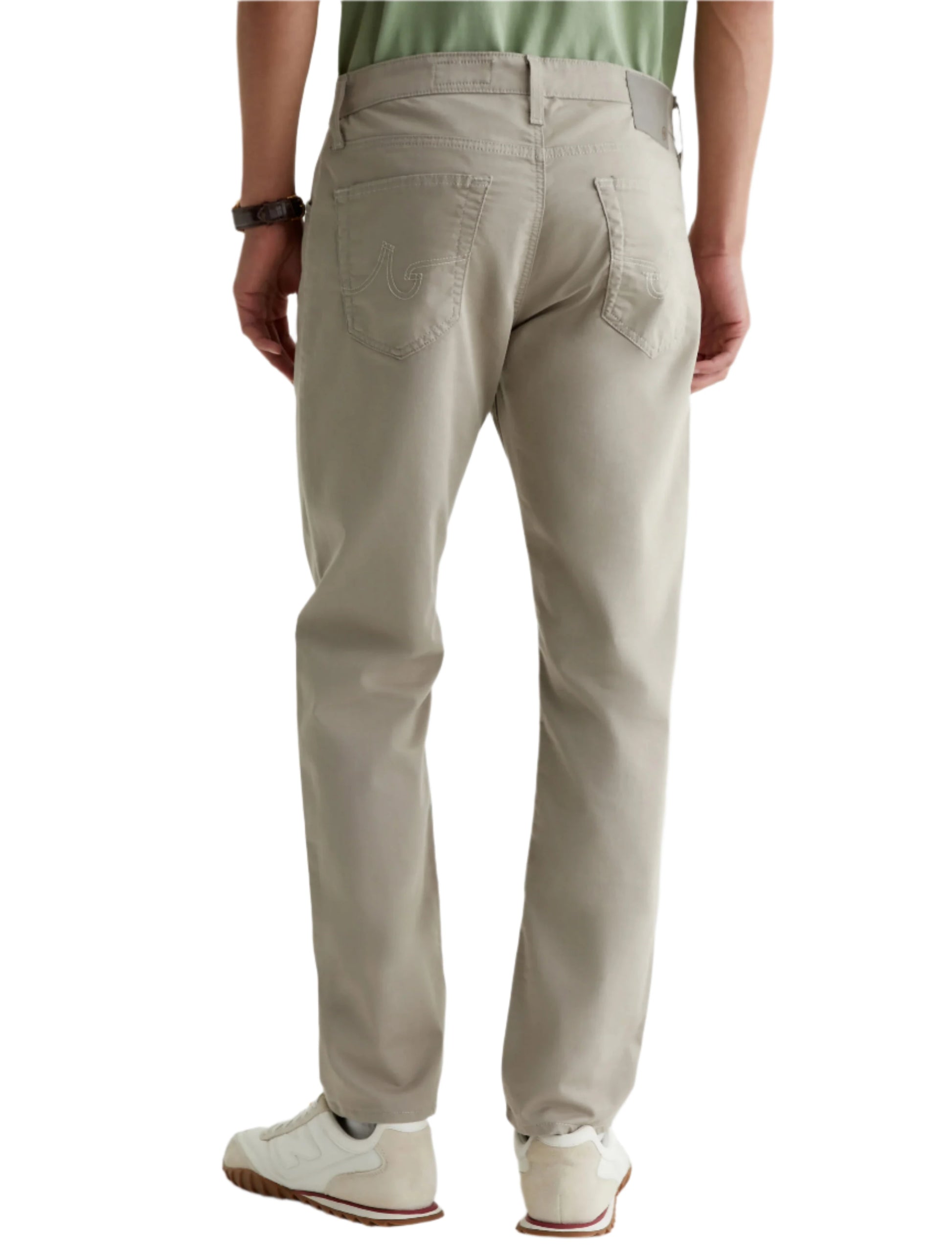 A person wearing AG Jeans Tellis Commuter Performance Airluxe Modern Slim jeans in light beige, paired with a green shirt, white sneakers, and a dark wristwatch, shown from the back.