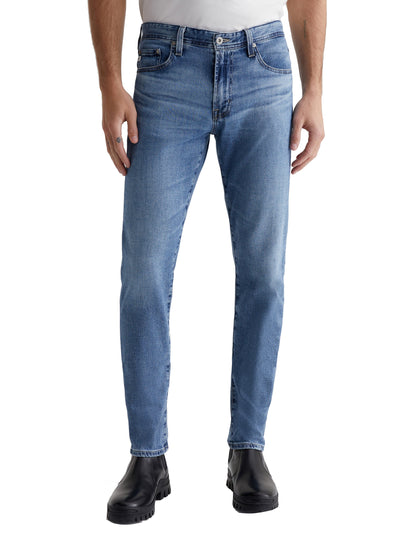 A person wears AG Jeans' Tellis Vapor Wash in light blue slim-fit, paired with black boots and stands against a plain white background.