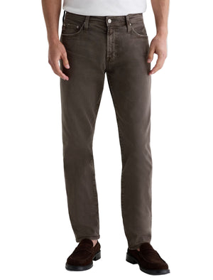 A person wears AG Jeans' Tellis Modern Slim Denim in brown, styled with a white shirt and brown loafers, standing against a plain background.