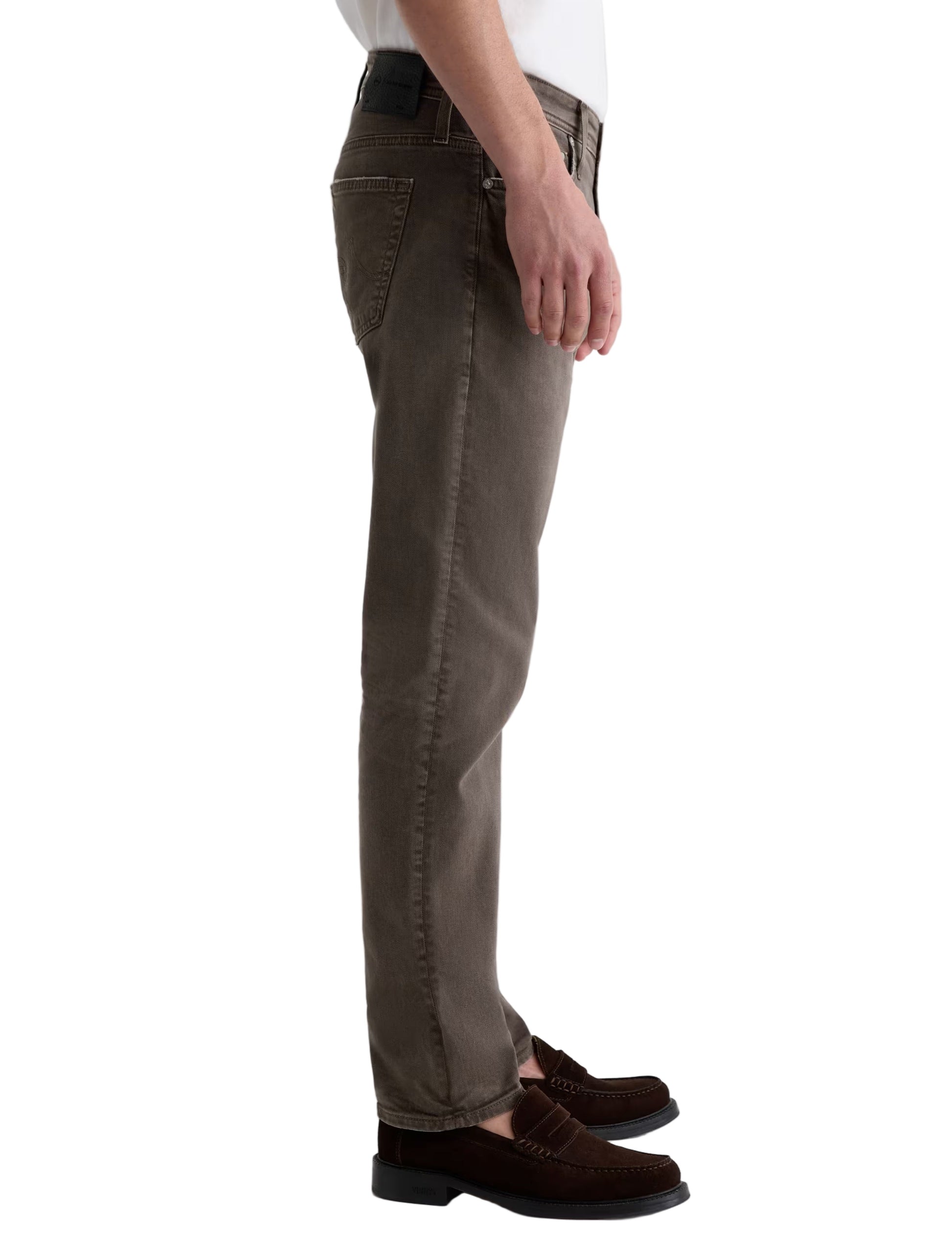 A person stands in side view against a plain background, wearing AG Jeans' Tellis Modern Slim Denim in Hyper Soft Bull Denim, paired with a white shirt and brown loafers.