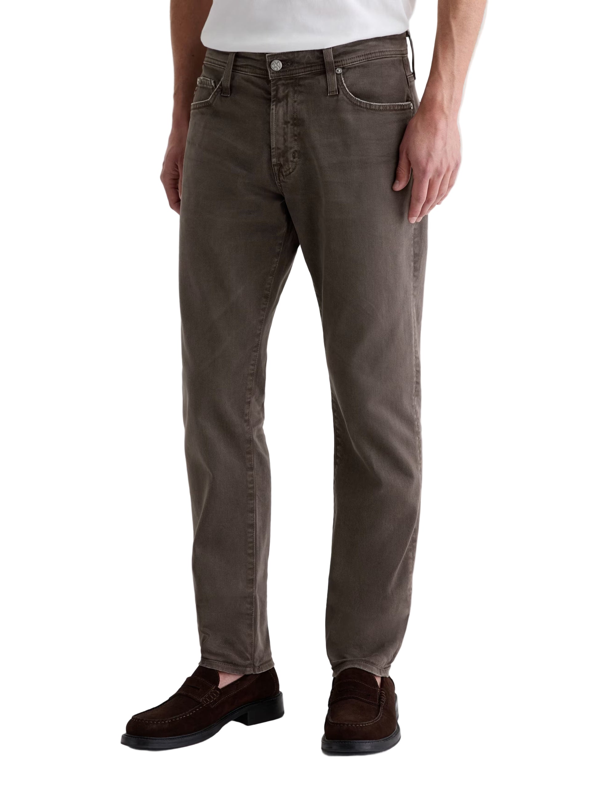 A man wearing AG Jeans' Tellis Modern Slim Denim, paired with a white shirt and brown loafers, stands against a plain background.