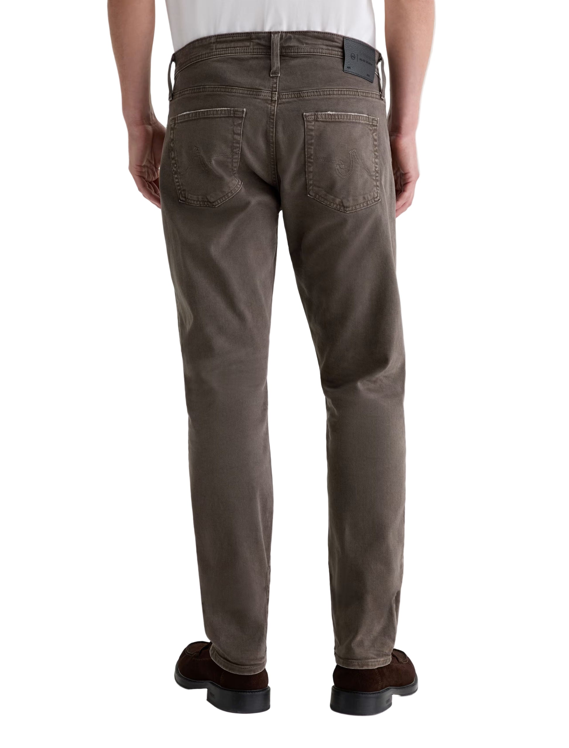 A person is shown from the back wearing brown Tellis Modern Slim Denim jeans by AG Jeans, a white shirt tucked in, and dark brown shoes.
