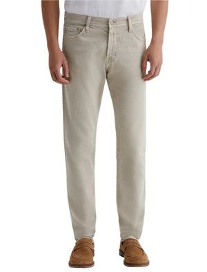 A person wearing AG Jeans Tellis AG-ed™ Modern Slim Denim in light beige, paired with a white shirt and brown suede shoes, is shown from shoulders to feet against a plain background.