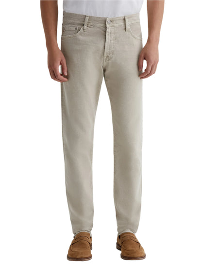 A person wearing AG Jeans Tellis AG-ed™ Modern Slim Denim in light beige, paired with a white shirt and brown suede shoes, is shown from shoulders to feet against a plain background.