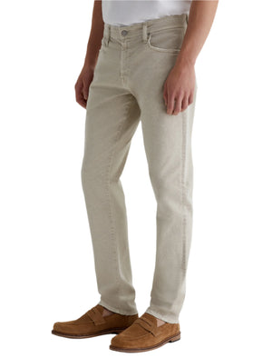 A person wears Tellis AG-ed™ Modern Slim Denim jeans by AG Jeans in light beige, paired with a white shirt and brown suede loafers, standing against a plain background.