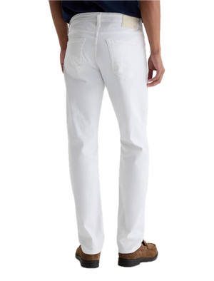 A person stands facing away, wearing AG Jeans’ Tellis Cloud Soft Denim™ white slim fit men’s jeans, styled with a navy shirt and brown suede shoes against a white background.