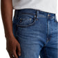 A person wearing a white t-shirt and AG Jeans Tellis Modern Slim Denim in medium blue, shown from waist to mid-thigh, with one hand by the pocket.