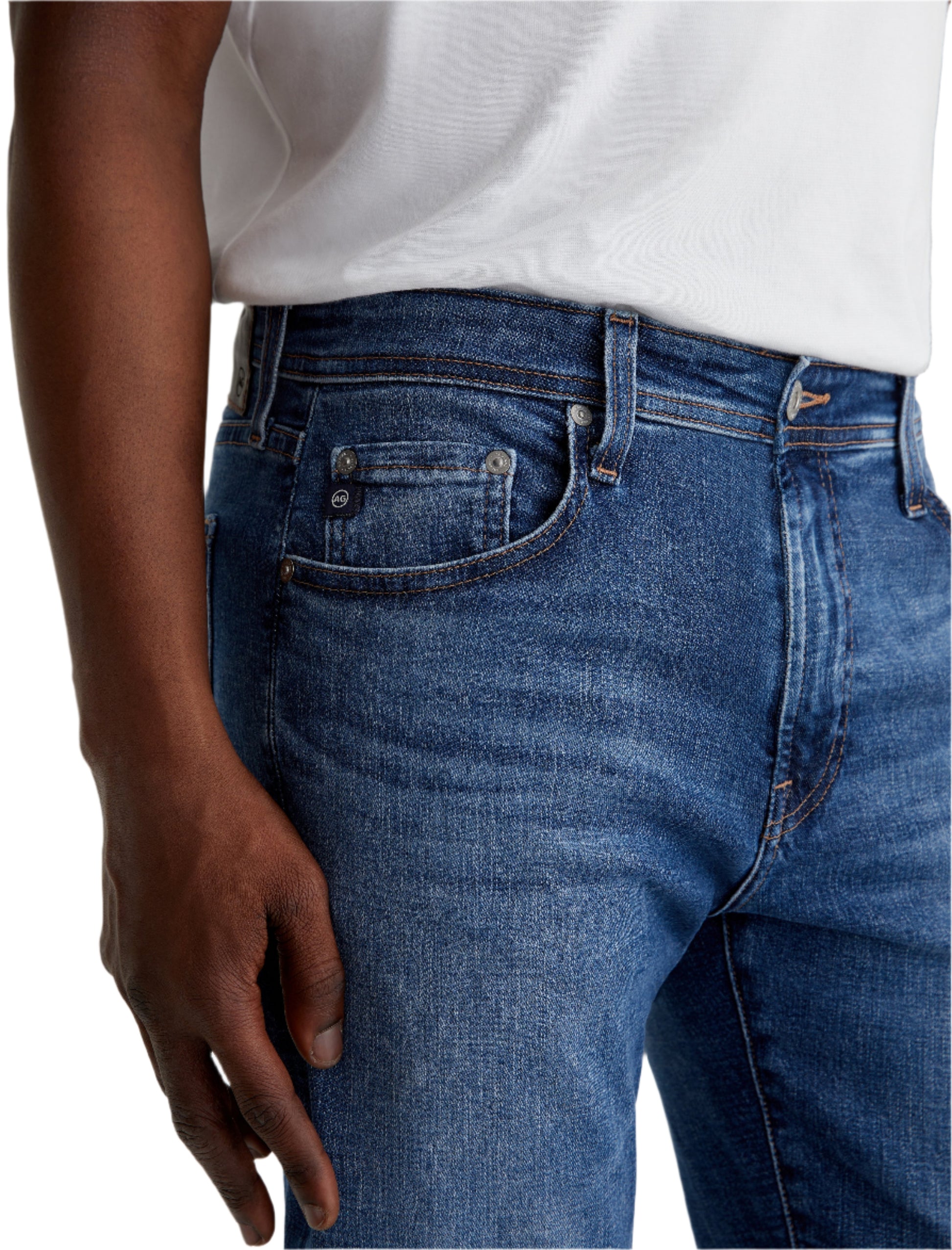 A person wearing a white t-shirt and AG Jeans Tellis Modern Slim Denim in medium blue, shown from waist to mid-thigh, with one hand by the pocket.