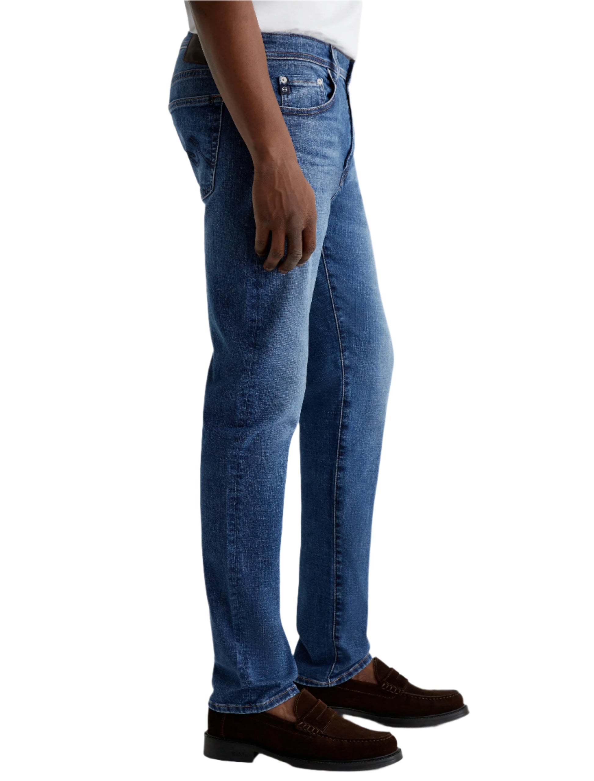 A man in AG Jeans Tellis Modern Slim Denim in medium blue, paired with a white shirt and brown loafers, stands with his arms relaxed at his sides.
