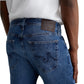 Model shows the back view of Tellis Modern Slim Denim by AG Jeans in medium blue, paired with a white shirt. Back pockets and brown leather waistband patch are visible.