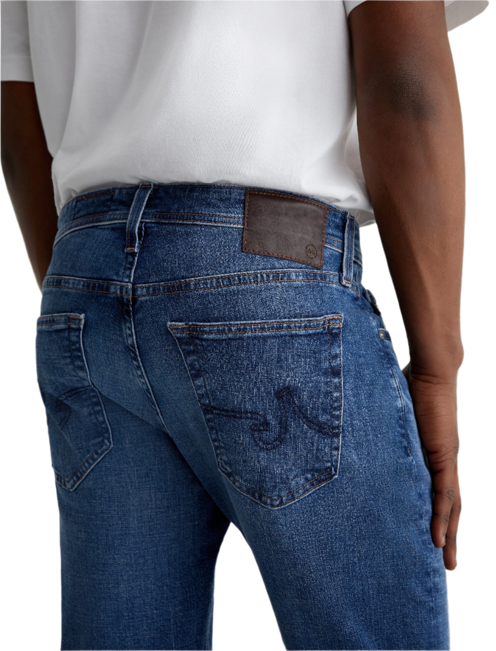Model shows the back view of Tellis Modern Slim Denim by AG Jeans in medium blue, paired with a white shirt. Back pockets and brown leather waistband patch are visible.
