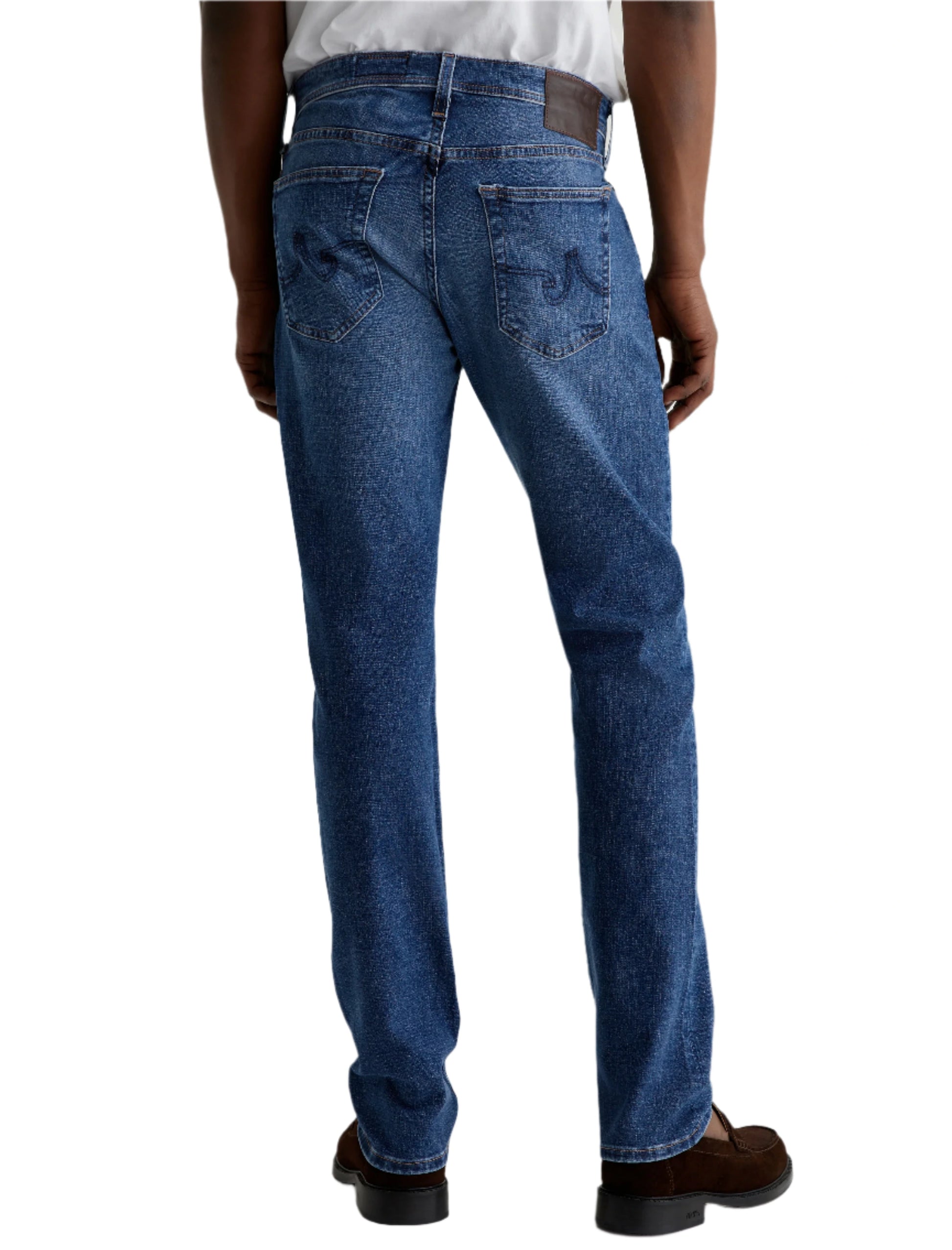 A person is shown from the back wearing AG Jeans Tellis Modern Slim Denim in medium blue, paired with a white shirt and brown shoes.