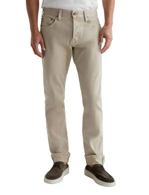 A person wearing AG Jeans Tellis Selvage khaki men's jeans in a modern slim fit, paired with a white shirt and brown suede shoes, stands against a plain white background.