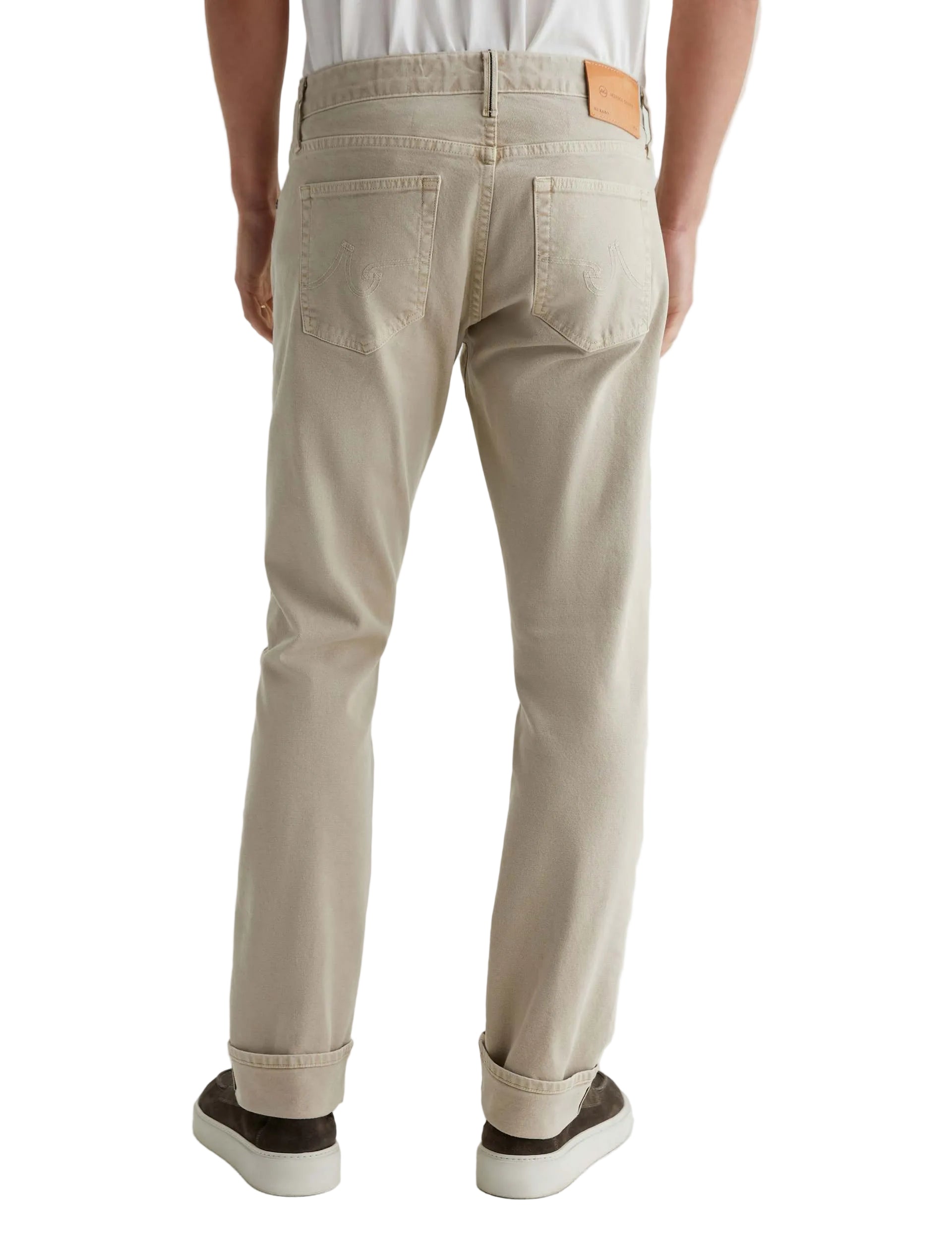 A person wearing AG Jeans Tellis Selvage khaki men's jeans, a white shirt, and dark sneakers stands with their back to the camera.