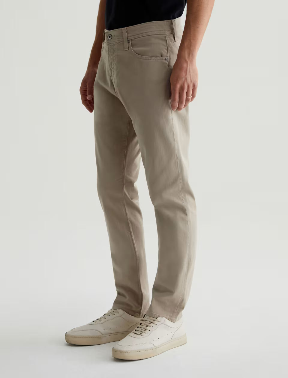 A person wearing AG Jeans Tellis Sueded Sateen Jeans in beige and white sneakers stands against a plain light background.