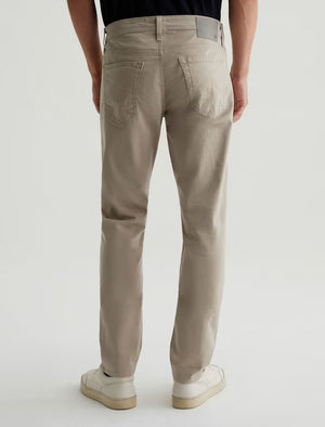 A person is shown from the back against a plain light background, wearing AG Jeans Tellis Sueded Sateen Jeans in beige paired with white sneakers.