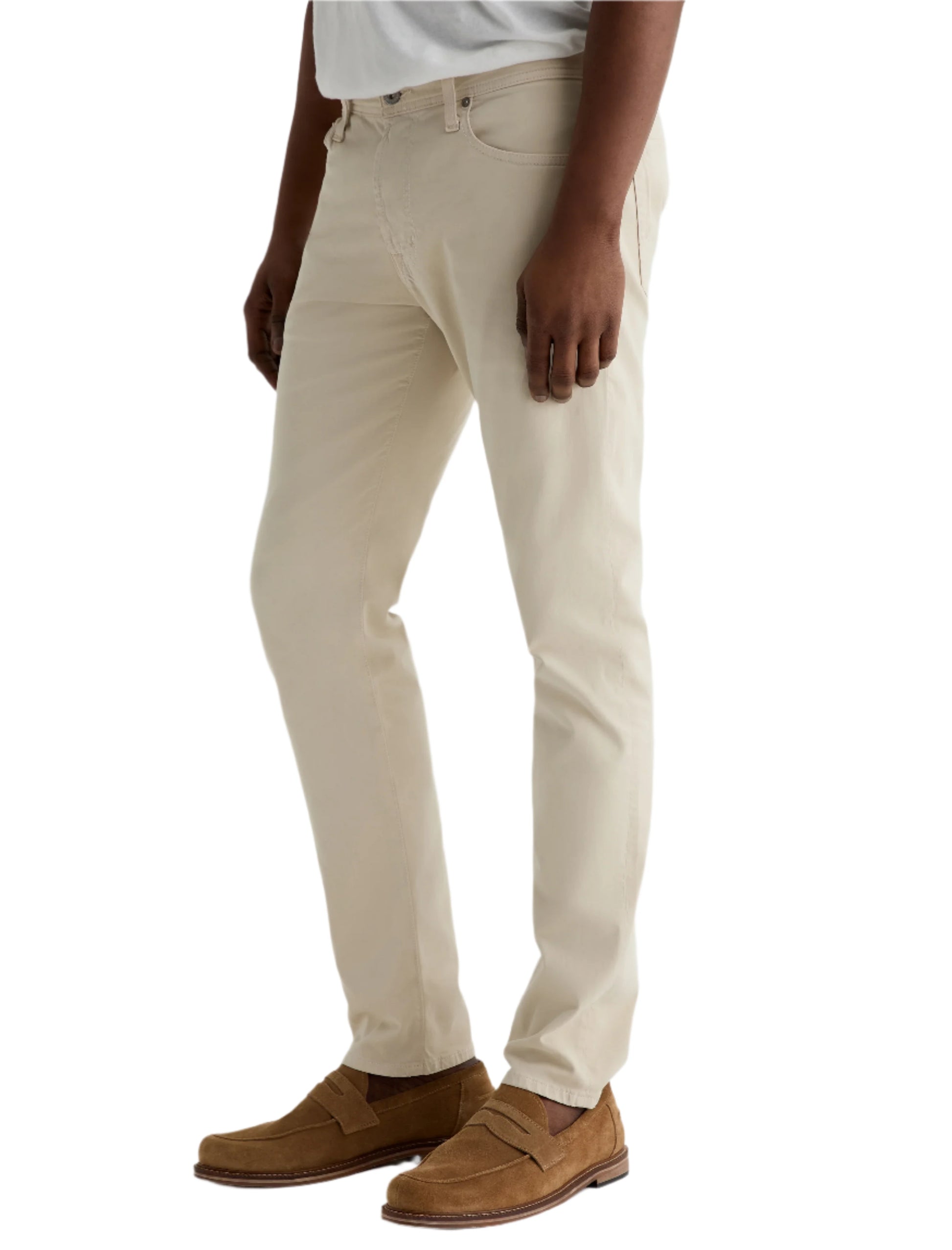 A person wearing light beige Tellis Modern Slim Sueded Stretch Sateen pants from AG Jeans, a light grey shirt, and brown suede loafers stands against a plain light background.
