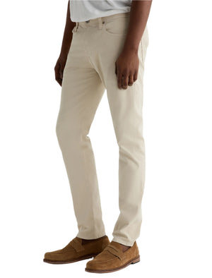 A person wearing light beige Tellis Modern Slim Sueded Stretch Sateen pants from AG Jeans, a light grey shirt, and brown suede loafers stands against a plain light background.