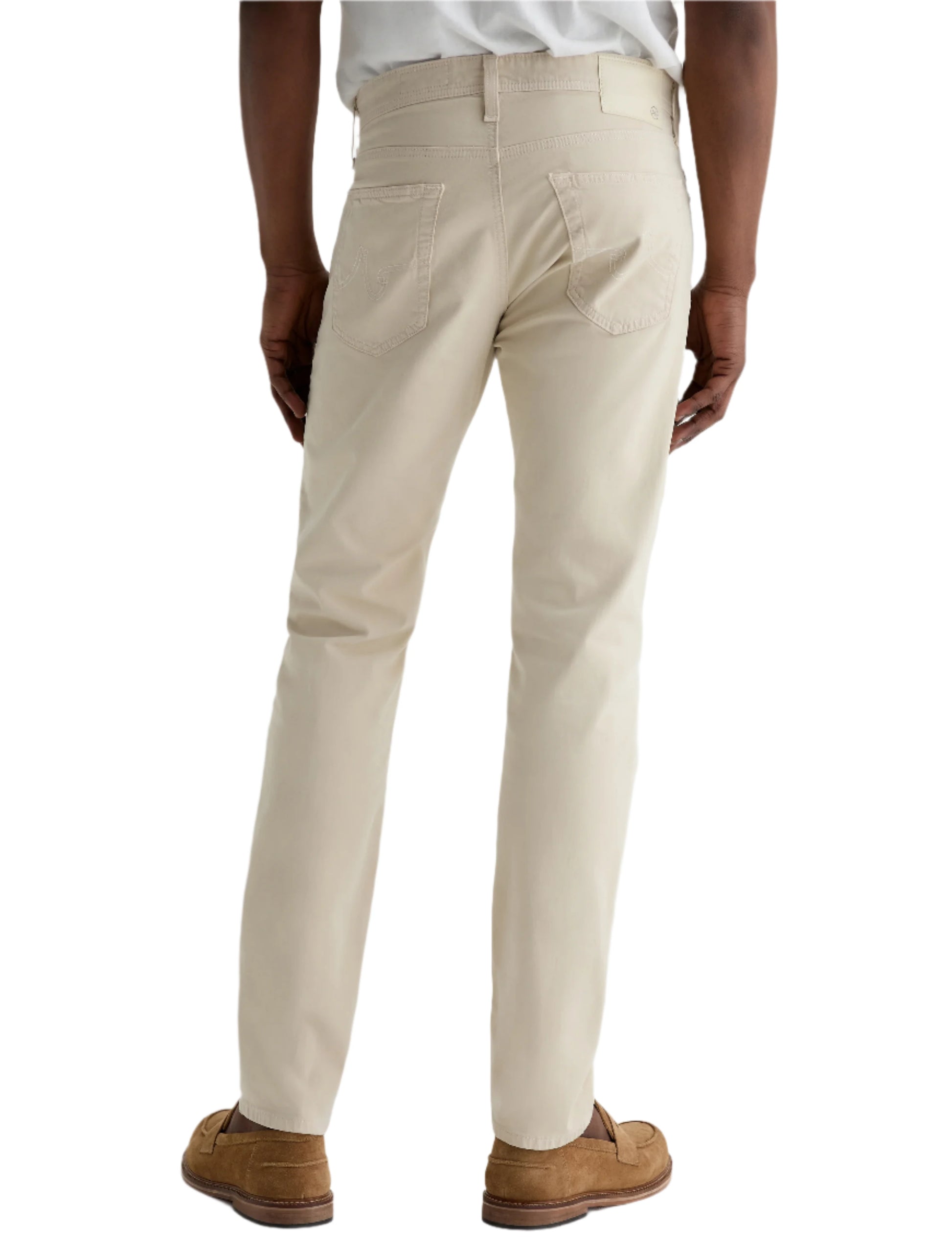 A person wearing AG Jeans' Tellis Modern Slim Sueded Stretch Sateen men's pants in light beige, paired with brown loafers, stands facing away from the camera against a plain background.