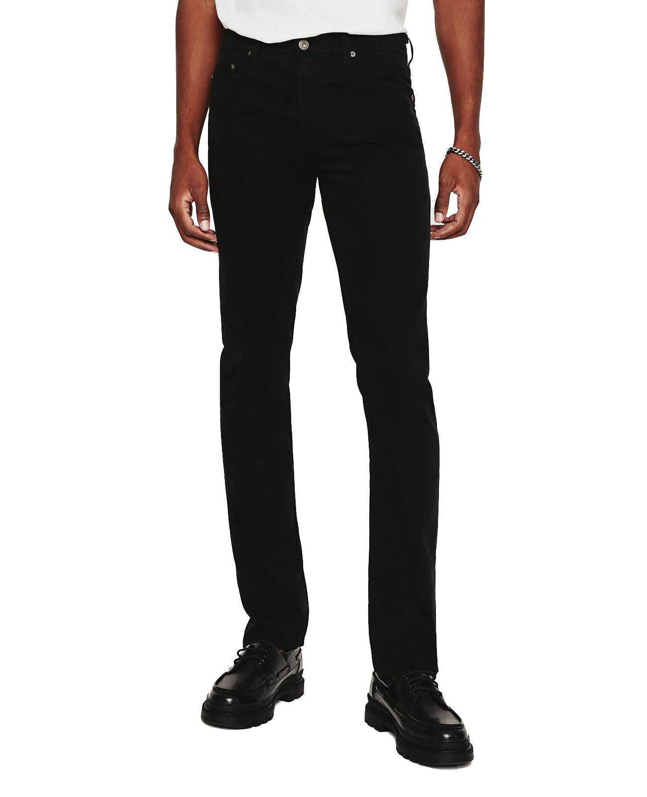 A person standing wearing comfortable AG Jeans Tellis Modern Slim black jeans and black shoes. The upper part of the body is not shown in the image.