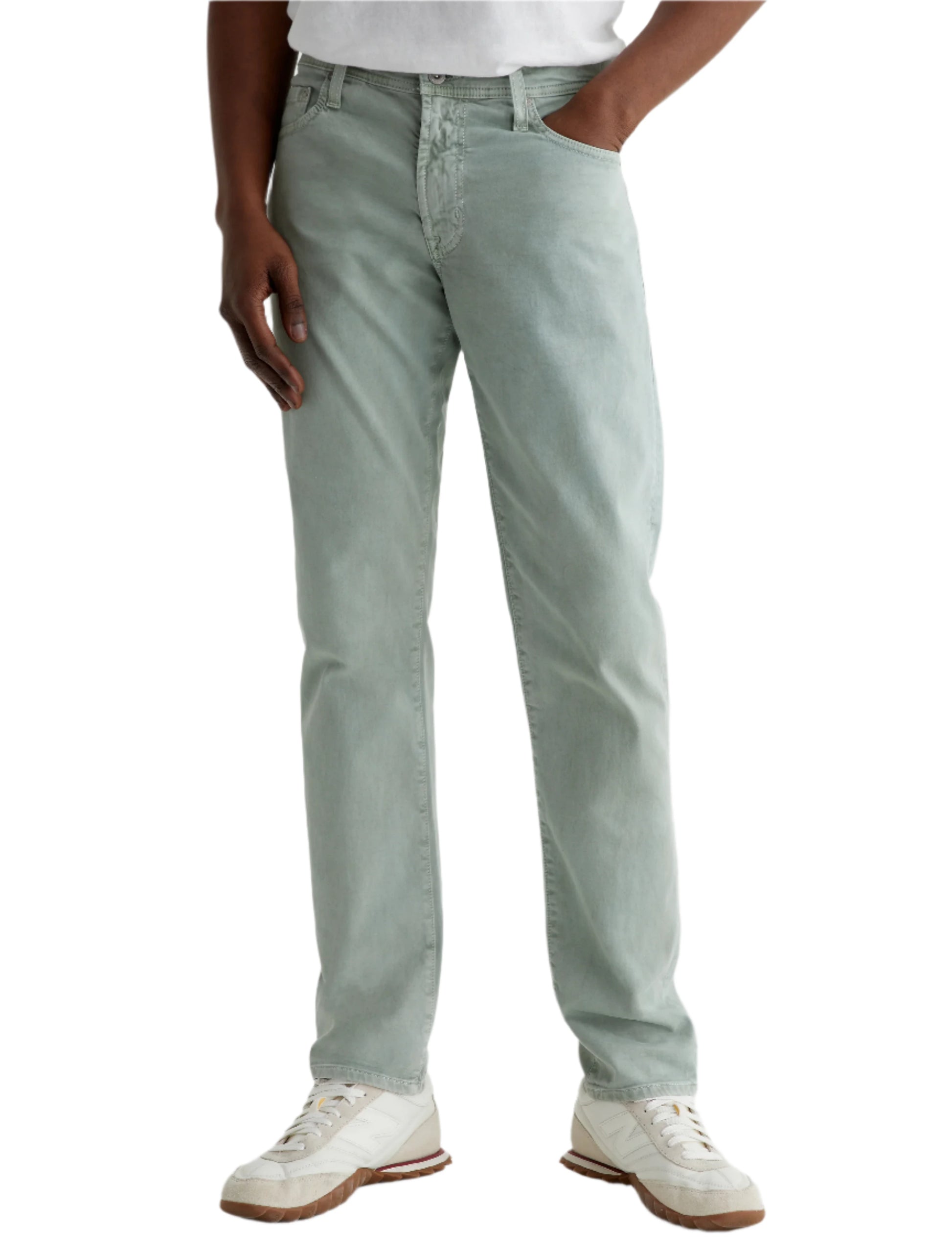 A person stands with one hand in their pocket, wearing AG Jeans Tellis Modern Slim Sueded Stretch Sateen light green men's pants, paired with a white shirt and white sneakers against a plain background.