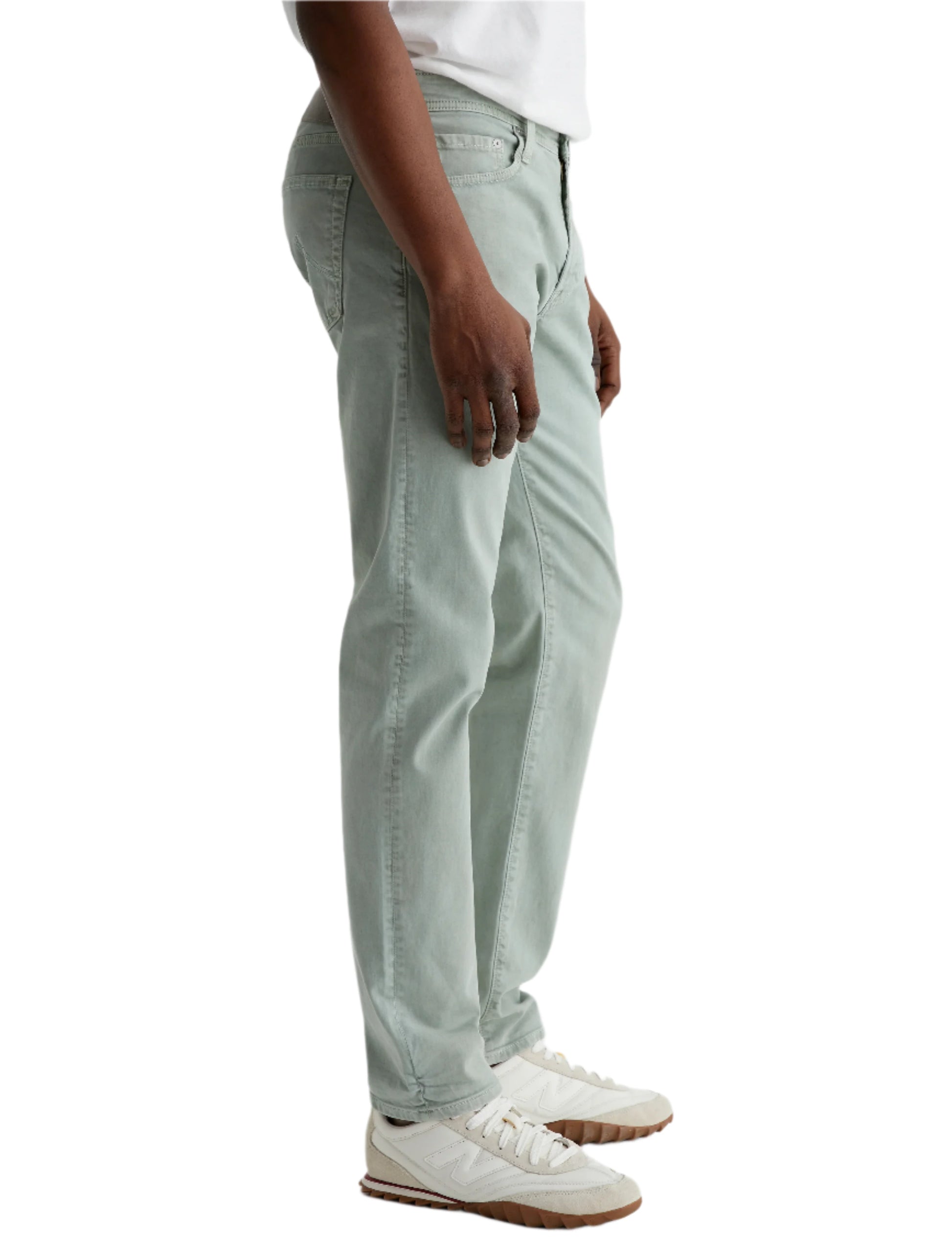 A person in side view wears AG Jeans' Tellis Modern Slim Sueded Stretch Sateen jeans in light green, paired with a white t-shirt and white sneakers with brown soles, set against a plain light background.