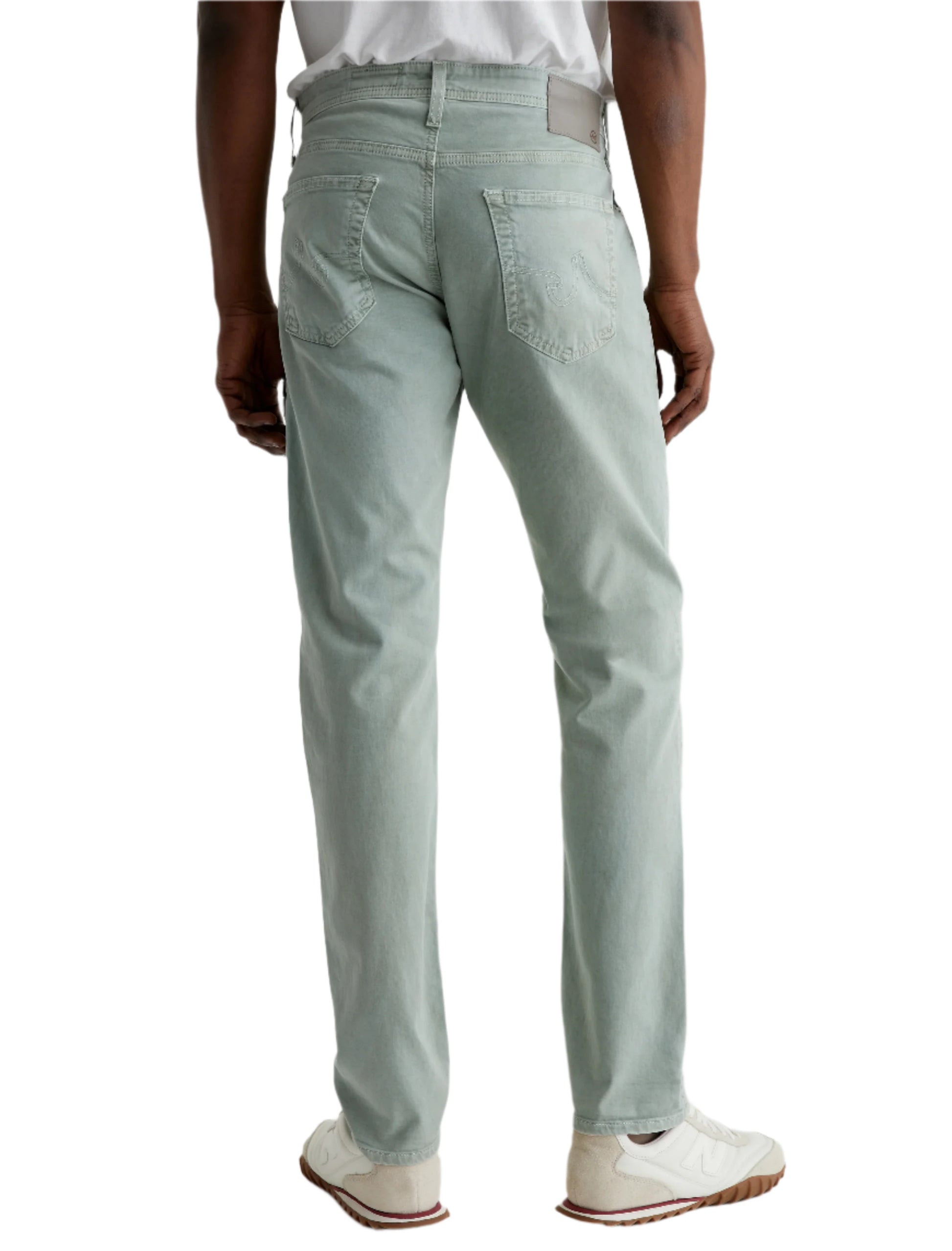 A person wearing AG Jeans' Tellis Modern Slim Sueded Stretch Sateen in light green, a white t-shirt, and white sneakers stands with their back to the camera against a plain background.