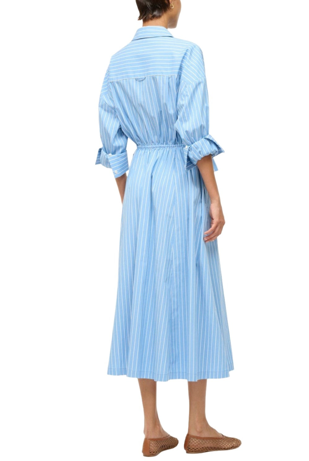 A person wearing the Staud Lisa Dress, a blue striped midi-length shirtdress with an elastic waist and long sleeves, stands with their back to the camera. They complement the outfit with brown woven shoes.