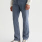 A person wearing AG Jeans’ Everett 7 Years Sulfur relaxed fit jeans in a grey-blue wash, paired with a light long-sleeve top and white sneakers with brown soles, stands against a plain background.