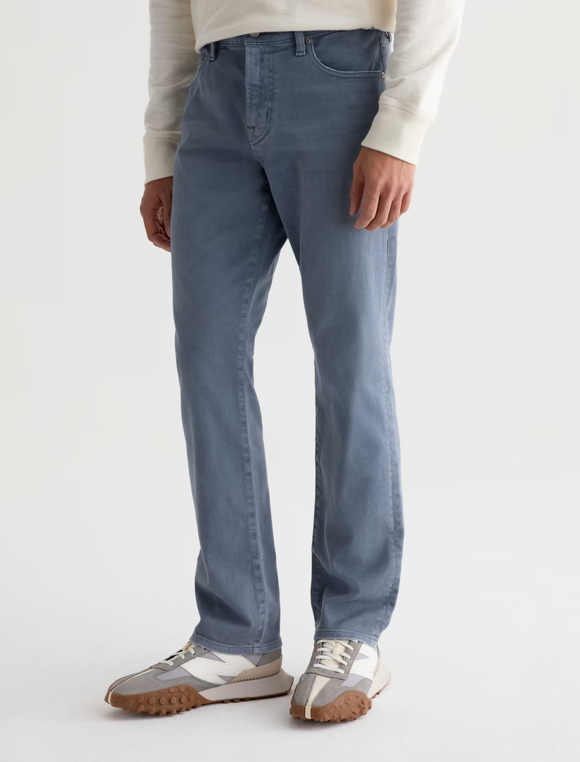 A person wearing AG Jeans’ Everett 7 Years Sulfur relaxed fit jeans in a grey-blue wash, paired with a light long-sleeve top and white sneakers with brown soles, stands against a plain background.