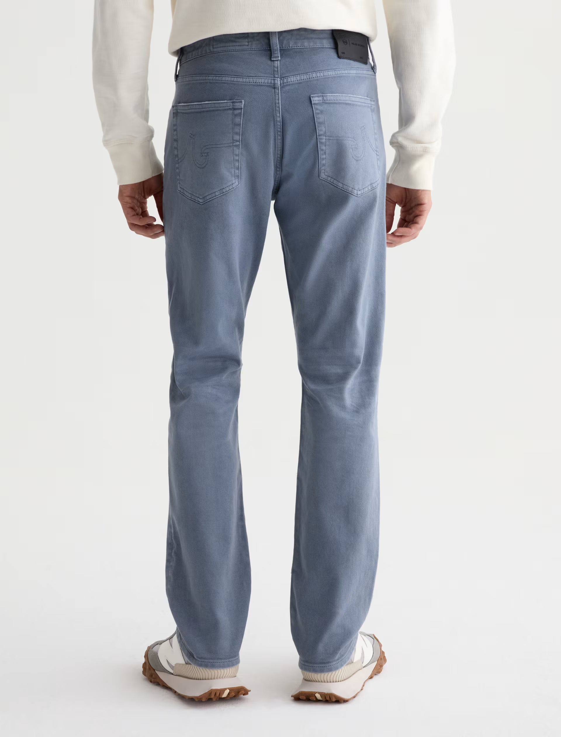 A man is shown from the back against a plain background wearing AG Jeans’ Everett 7 Years Sulfur slim straight grey-blue jeans, paired with a white long-sleeve shirt and white sneakers.