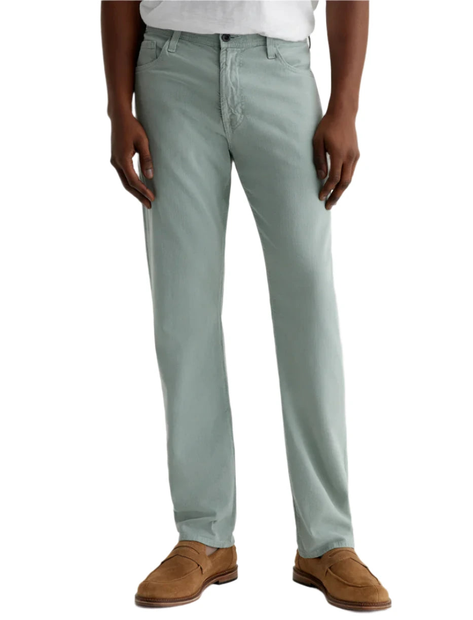 A person in AG Jeans' Everett Slim Straight Leg men's pants in light green, paired with a white shirt and brown suede shoes, stands against a plain background.