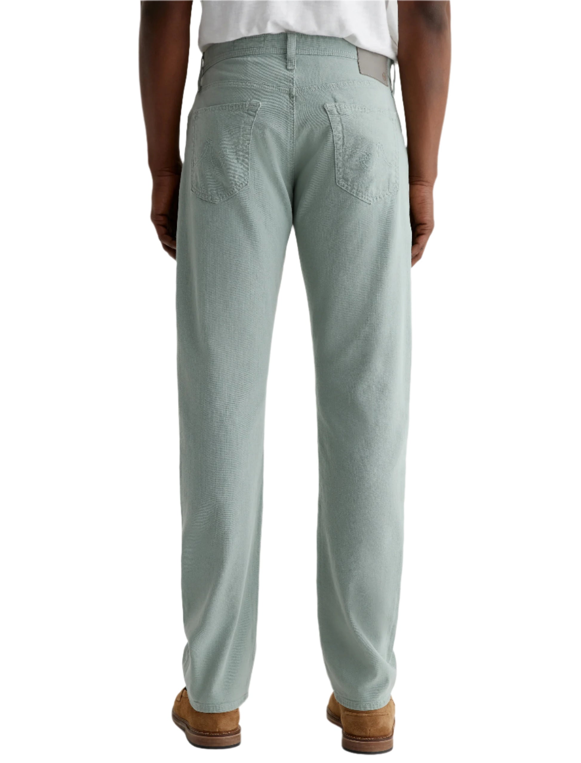 A person is shown from the back, standing against a plain background, wearing AG Jeans Everett Slim Straight Leg men's pants in light green, paired with a white shirt and brown shoes.