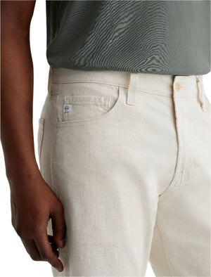 A person is shown from waist to mid-thigh wearing AG Jeans' Everett Slim Straight Leg in off-white and a green textured shirt, with one hand relaxed by their side.