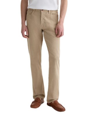 A person stands against a plain white background wearing AG Jeans' Everett Slim Straight beige jeans, a white shirt, and brown loafers.